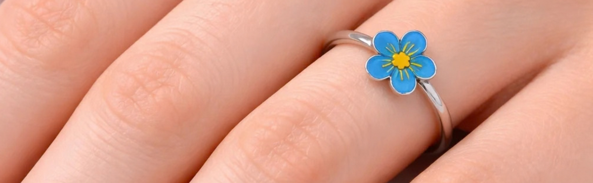 A close-up of a hand wearing a silver ring with a small blue flower charm featuring a yellow center.